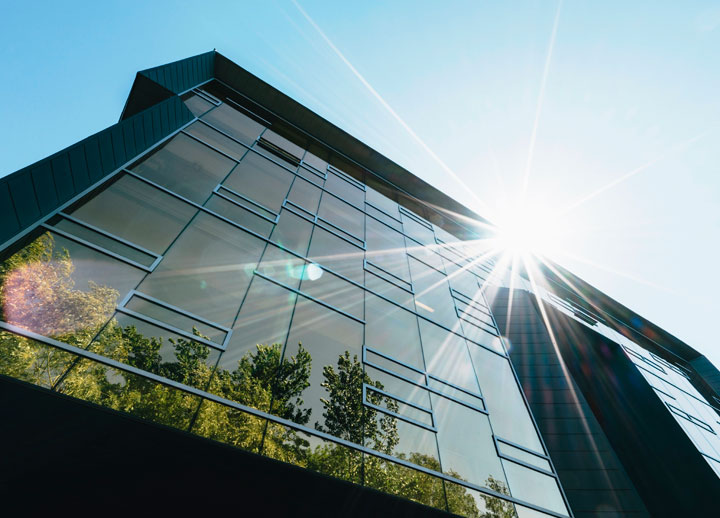 Imatge en contrapicat d'un edifici amb la façana de vidre i alumini, reflex dels arbres i rajos de sol en contrast.